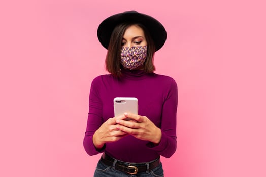 Stylish woman in protective  mask and black hat using mobile phone , standing on pink background. photo