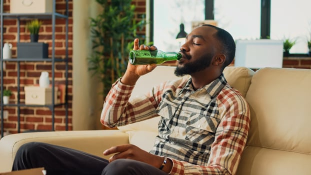 African american person eating burger and fries on sofa, having dinner with fast food and bottle of alcohol at home. Young man feeling happy watching action movie or show, having fun. photo