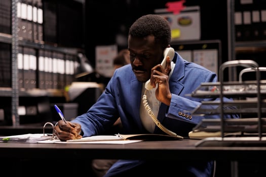 African american police officer talking with remote detective using landline phone, discussing confidential clue. Private inspector analyzing crime scene evidence trying to solve case in arhive room photo