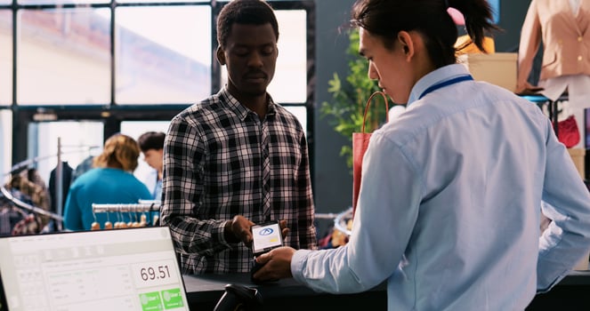 African american client buying stylish clothes, paying for trendy clothes using mobile phone nfc on pos terminal in modern boutique. Shopper man shopping for elegant merchandise. Fashion concept photo