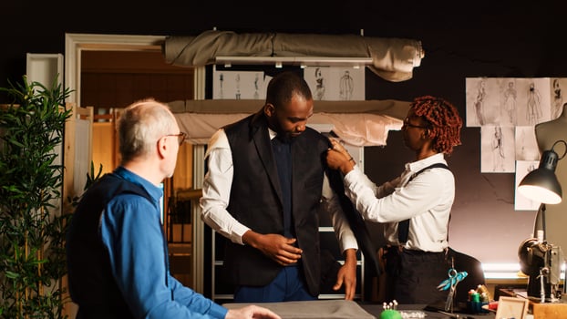 Young customer trying on suit jacket in atelier, making sure it fits with the right measurements. Experienced tailor and assistant designing custom made blazer, fitting in workshop. photo