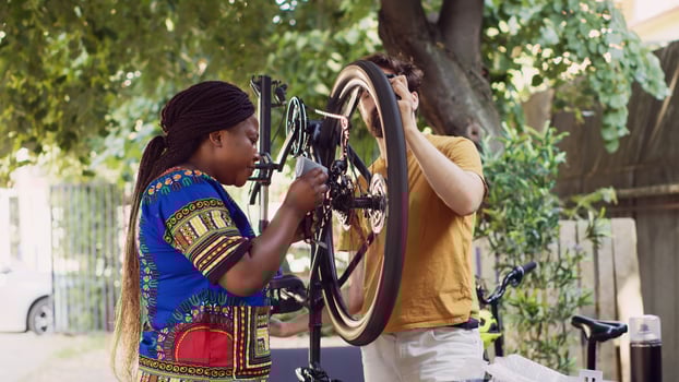 Active motivated interracial couple outside adjusting bike wheel with professional equipment. Young black woman grabbing specialized tools to assist caucasian man in fixing bicycle. photo