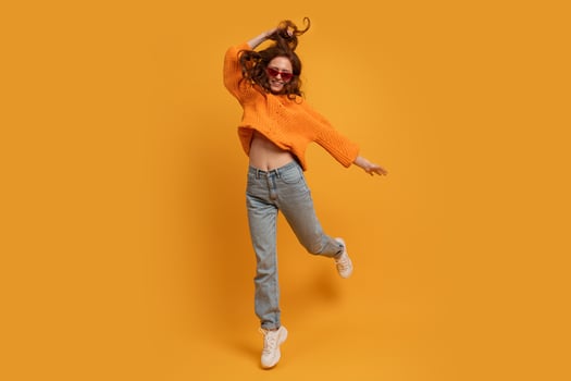 Laughing stylish red head  woman in orange sweater and blue jeans  jumping  over yellow  background.  Full lenght. photo