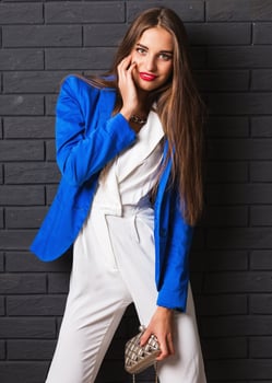 Fashionable studio image of   stylish attractive young woman in casual white costume and blue jacket holding luxury purse posing on black urban brick wall background. photo
