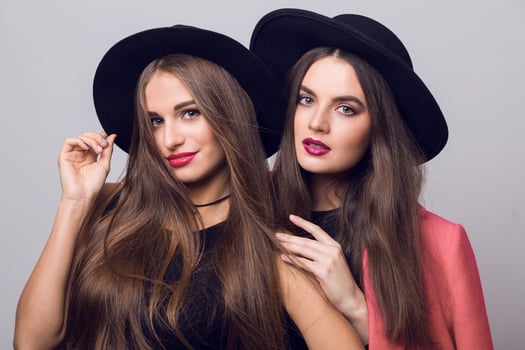 Close up beauty  fashion image of two girls, sisters, posing on grey background, hugging, smiling. Wearing stylish pink  coat and black hat, short top and evening dress. photo
