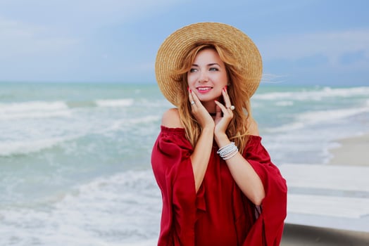 Close up portrait of smiling pretty woman in straw hat and trendy red summer dress posing near ocean waves. Windy  and cloudy weather. photo