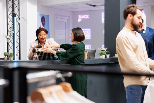 Black Friday and retail industry. Young Asian woman customer standing at clothing store checkout counter giving clothes to friendly cashier, buying garment at discounted prices during seasonal sales photo