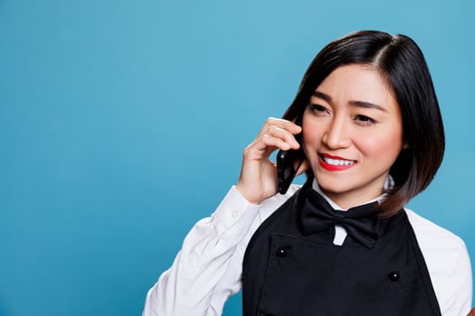 Hotel smiling cheerful asian woman receptionist wearing uniform speaking on smartphone. Joyful waitress answering call and chatting with catering service customer on mobile phone photo