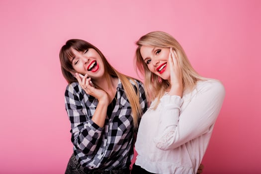 Brunette woman with surprise face talking by mobile phone. Her best friend  laughing and having fun. Pink background. Studio shot. photo