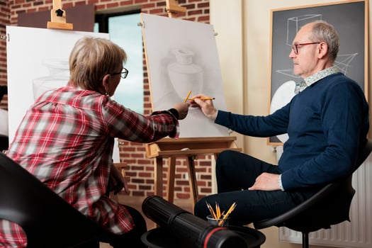 Retired couple talking discussing artwork while learning new skill. Senior man and woman sitting at easels learn to draw, attending group art class together. Creative activities to do with family photo