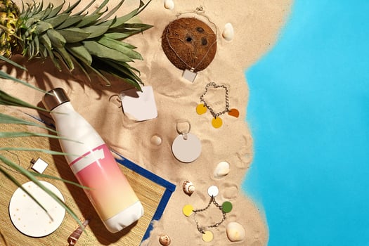 Jewelry, cosmetic bottle with no label, shells, coconut, pineapple, palm twig and keychains on sand and blue background. Close up, copy space photo