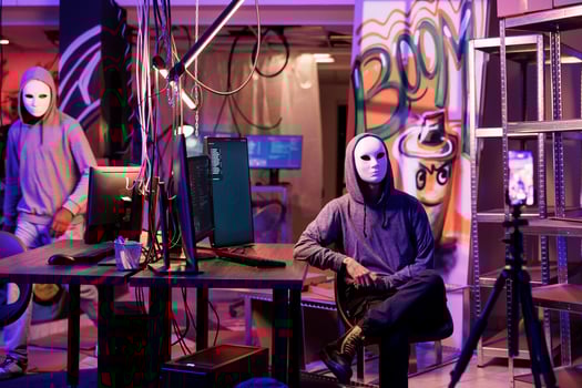 Anonymous hacker in mask recording threat video on smartphone camera in abandoned warehouse. Cybercriminals breaking law while streaming internet fraud and cyberattack on mobile phone photo