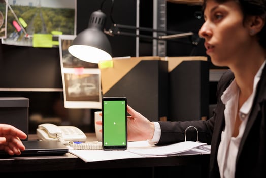 Private detective holding smartphone with green screen mock up display, discussing criminology report with police officer. Investigators working overtime in evidence room. Phone with isolated template photo