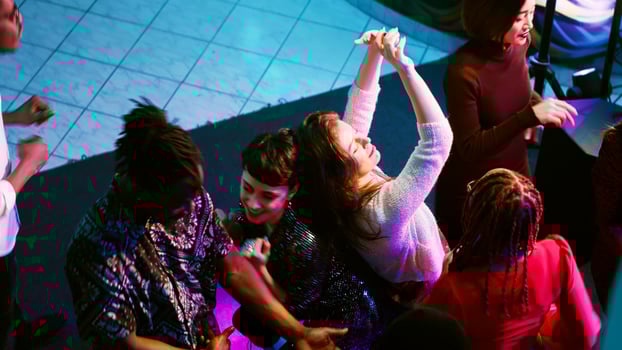 Happy persons clubbing together at night, listening to modern music from DJ station panel on stage. Group of friends partying and dancing in nightclub, spotlights event. Handheld shot. photo