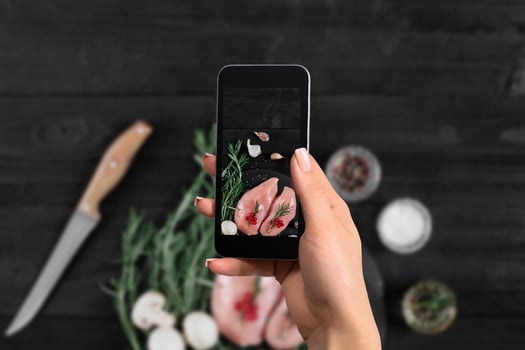Female hand with a smartphone makes a photo raw chicken breast on black background, with fresh mushrooms, garlic and herbs.