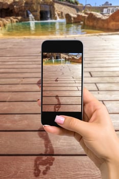 Closely image of female hands holding mobile phone with photo camera mode on the screen. Cropped image of footprints on the wooden floor behind it swimming pool