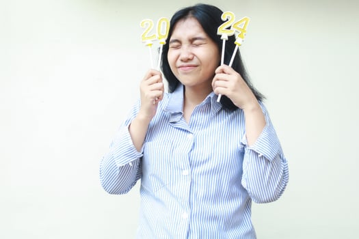 happy and excited asian young woman holding golden candles number 2024 stick on her head with eyes closed to celebrate new years eve wear blu stripes shirt isolated on white photo
