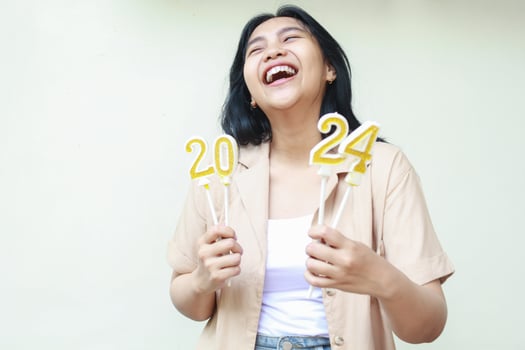 portrait of happy asian young woman hipster laughing with open mouth and looking aside and up while holding 2024 figure candles wearing stylish brown outer isolated on white background photo