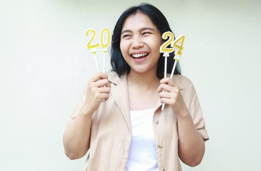 laughing asian young woman holding 2024 gold candles and looking aside with toothy smile wearing brown casual outer isolated on white background photo