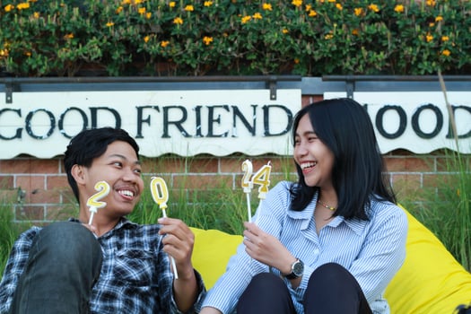 happy asian couple teen looking to each other holding 20 24 glittering candle and sitting on yellow bean bag in house yard meadow with flower background in eveningto celebrate new year photo