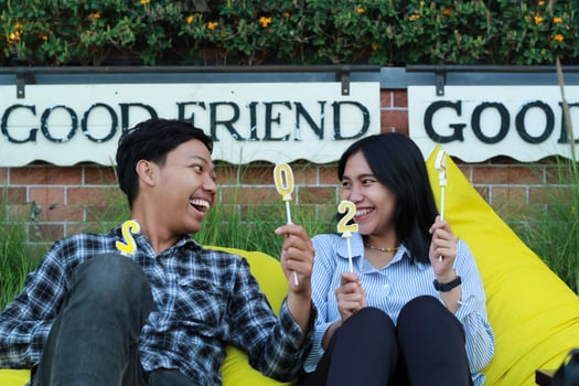 happy asian couple laughing looking to each other with raised hand holding candle number 20 24 sitting on the yellow couch or bean bag in the house backyard with brick wall and flower grass background photo