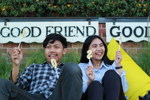 happy asian couple teenager raising number 20 24 golden candles while sitting on the yellow bean bag in back yard with brick wall background, look away photo