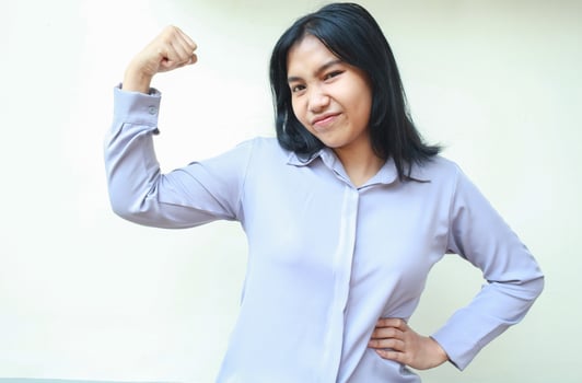 carefree powerful asian young business woman raising arm excited showing biceps strenght with hands on waist wearing formal shirt standing over white background, looking at camera photo