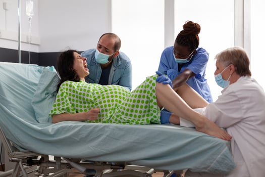 Diverse nurse and medic helping with childbirth in hospital ward. Mother lying in bed having painful contractions giving birth to baby while father standing beside her comforting in maternity clinic photo