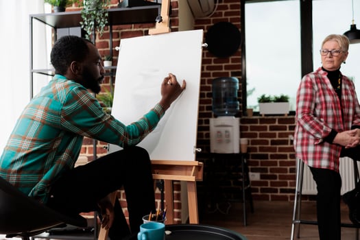Young focused African American guy learning to draw with teacher, male student sitting at easel in classroom sketching simple object with pencil, taking up art classes. Drawing and selfexpression photo