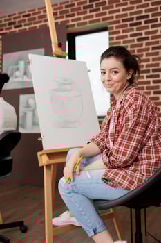 Happy young woman smiling at camera while sitting at easel during fun and creative art class, learning basics of drawing, trying something new. Student girl enjoying arts activities photo