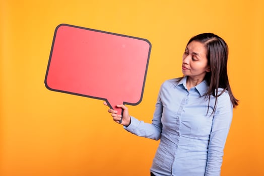 Asian young adult holding empty red speech bubble with copy space, advertising beauty product. Woman standing in studio over yellow background, looking at blank dialog frame with place for text photo