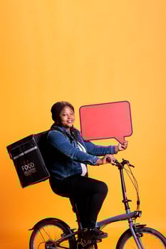 Smiling african american restaurant employee holding red speech bubble promoting takeaway food service while delivering fast food order to client using bike as transportation. Takeout concept photo