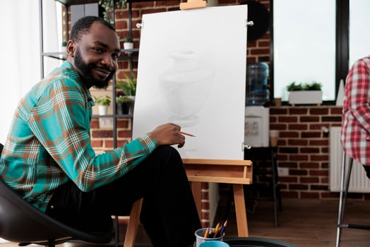 Art activities and wellbeing. Portrait of happy smiling African American guy during drawing lesson, young black man sitting at easel looking at camera, feeling inspired during sketching class photo