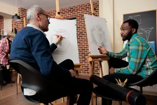 Two diverse men art school students talking during lesson, sitting at easels discussing artwork during creative group class, friends learning basics of drawing and sketching together photo