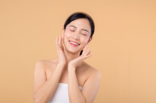 Beautiful girl asian model touching fresh glowing hydrated facial skin on beige background closeup. Beauty face young woman with natural makeup and healthy skin portrait. Skin care concept photo