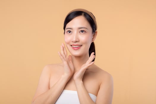 Beautiful girl asian model touching fresh glowing hydrated facial skin on beige background closeup. Beauty face young woman with natural makeup and healthy skin portrait. Skin care concept photo