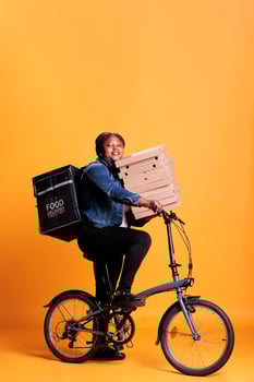Restaurant worker riding bike while carrying stack of pizza delivering takeaway food meal to customer Pizzeria employee standing in studio with yellow background. Food service and transportation photo