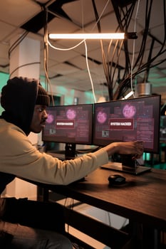 African american spy looking at hacking alert on multiple screens, planning security breach to steal personal information or passwords. Young adult using trojan virus and computer malware. photo