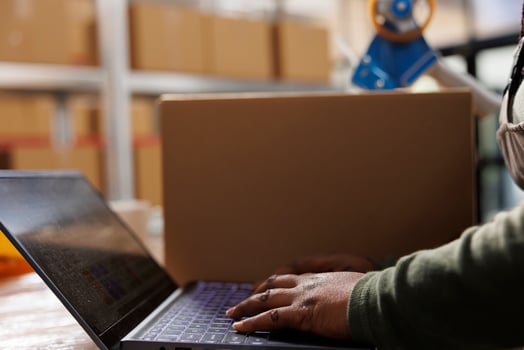 Stockroom worker checking goods quality control report on laptop computer before start working at customers order in warehouse. African american employee preparing packages for delivey. Close up photo