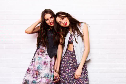 Close up   portrait of two pretty hipster  sisters  wearing boho colorful dress, stylish jeans jacket  and  baubles. Girls smile, have fun against  urban white wall. photo