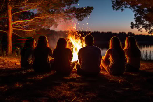 Silhouette of a group of young people sitting around a bonfire in the forest at sunset, Friends sitting in front of a bonfire, top section cropped, no visible faces, AI Generated photo