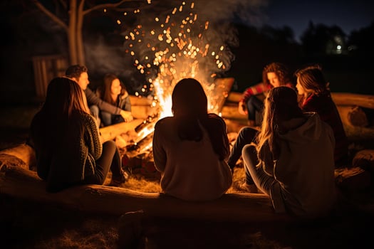Group of young friends sitting near bonfire at night. Camping concept, Friends sitting in front of a bonfire, top section cropped, no visible faces, AI Generated photo