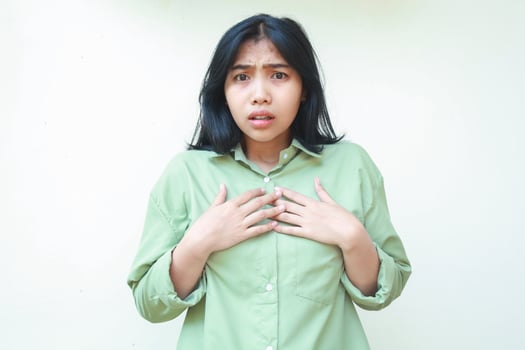 shocked asian woman with dark hair looks scared with hands touching chest wearing green oversized shirt isolated on white background photo