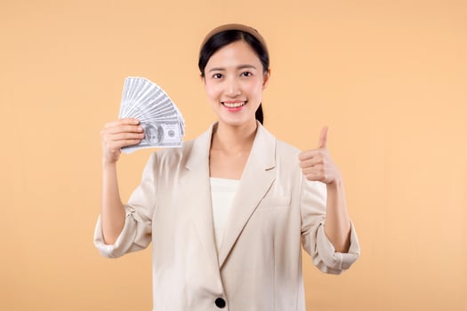 portrait of happy successful confident young asian business woman wearing white jacket holding cash money dollars standing over beige background. millionaire business, shopping concept. photo