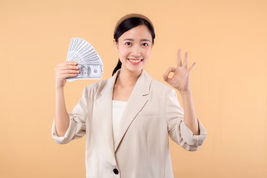 portrait of happy successful confident young asian business woman wearing white jacket holding cash money dollars standing over beige background. millionaire business, shopping concept. photo