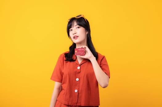 Portrait young beautiful asian woman happy smile dressed in orange clothes showing credit card isolated on yellow background. Pay and purchase shopping payment concept. photo