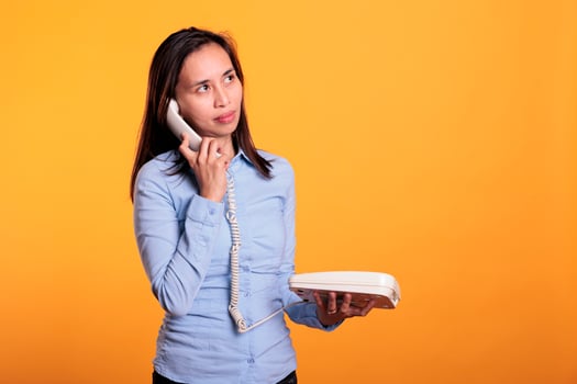 Asian woman talking at landline phone with remote worker, chatting on retro telephone line with cord in studio over yellow background. Cheerful smiling young adult enjoying lifestyle discussin photo