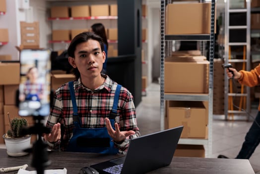 Postal service shipment manager filming order tracking operation on mobile phone camera. Warehouse asian employee sitting at desk in storage room and advertising goods for social media photo