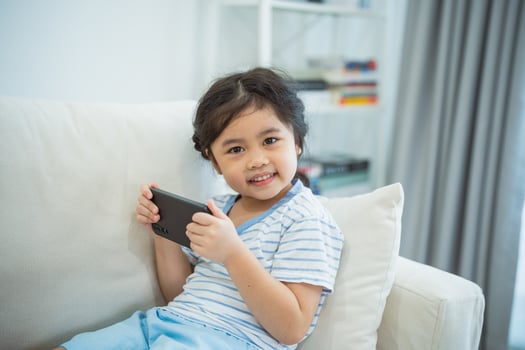 Asian child girl looking using and touch mobile phone screen on couch sofa. Baby smiling funny time to use mobile phone. Too much screen time. Cute girl watching videos while tv, Internet addiction. photo