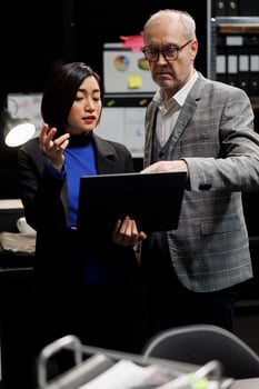 Elderly detective and asian private investigator team checking criminology suspect case file details on laptop. Teamworking investigation coworkers in archival agency depository office photo